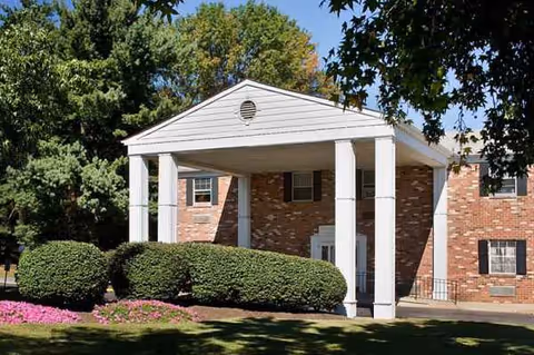 Brick senior living facility with white, columned front entrance under a peaked portico, surrounded by shrubs.