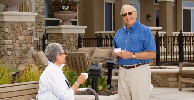 An elderly man and woman enjoying coffee together outside in a courtyard area with stone walls and outdoor seating at a senior living facility.
