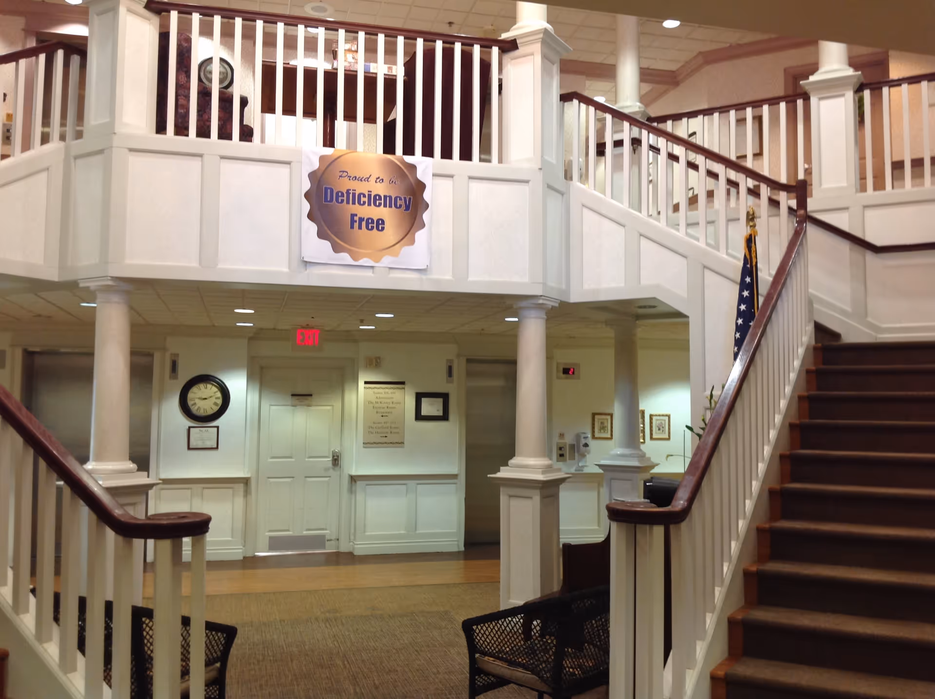 Interior view of a healthcare facility lobby with a staircase on the right, white railings, columns, and a sign that reads 'Proud to be Deficiency Free' hanging above the entrance to a hallway with an elevator and a clock on the wall.
