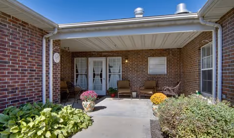 Covered brick patio with chairs, potted flowers, and double doors leading into the building.