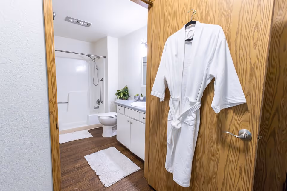 View into a bathroom with a white bathtub and shower combination, a toilet, and a sink with a countertop. A white bathrobe hangs on the wooden door, and two white bath mats are on the wooden floor. There are small plants on the countertop next to the sink.