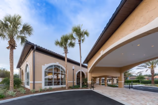 Exterior view of a senior living facility with beige stucco walls, large arched windows, and a covered entrance. There are several palm trees and landscaped greenery around the building, with a paved driveway and parking area in front.