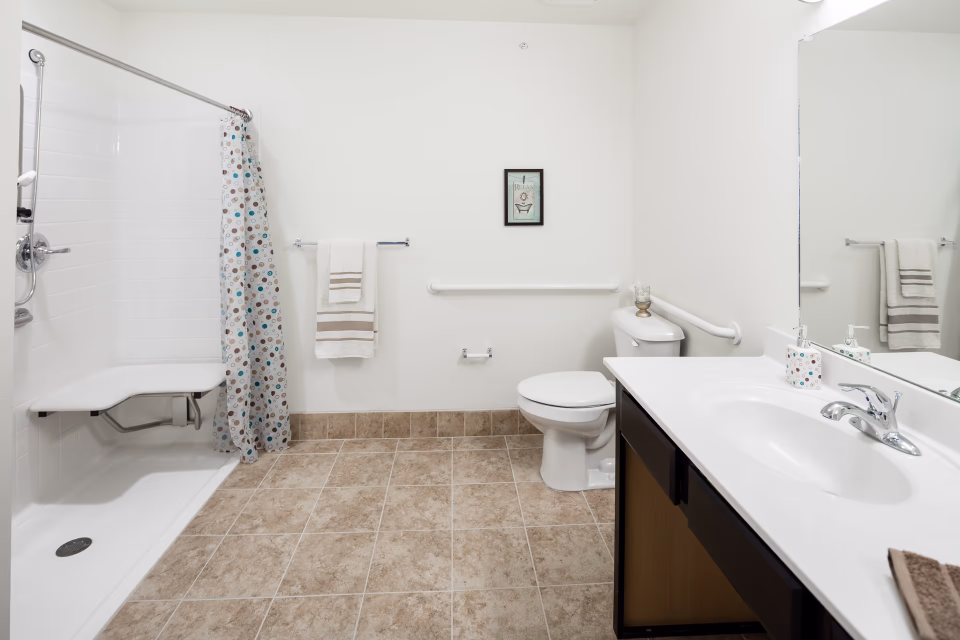 A clean and accessible bathroom with a walk-in shower featuring a fold-down seat and a shower curtain with circular patterns. There is a white toilet with grab bars on the wall behind and beside it. A towel rack with a white towel is mounted on the wall, and a framed picture hangs above the grab bar. The bathroom has beige tiled flooring and a white countertop with a sink and a soap dispenser. A large mirror is mounted above the sink.