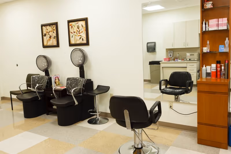 Interior view of a hair salon area in a senior living facility with two black hair drying chairs, a black styling chair in front of a large mirror, a wooden shelving unit with hair products, and a small sink area in the background.