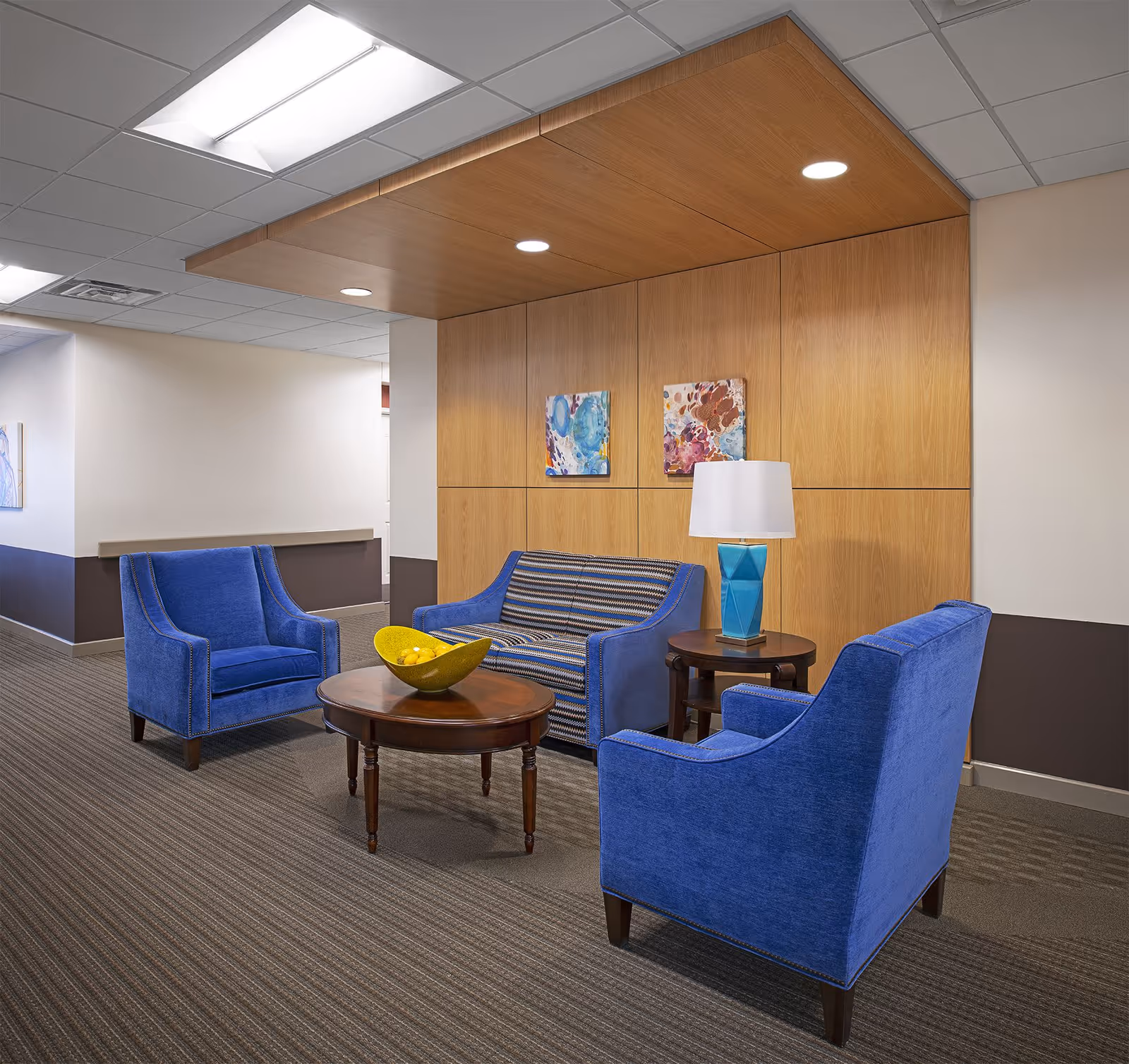 A cozy seating area in a senior living facility with two blue armchairs and a blue patterned loveseat arranged around a wooden coffee table with a yellow bowl of lemons. There is a small round side table with a blue lamp, and two colorful abstract paintings hang on a wooden paneled wall. The ceiling has recessed lighting and a wood accent panel.