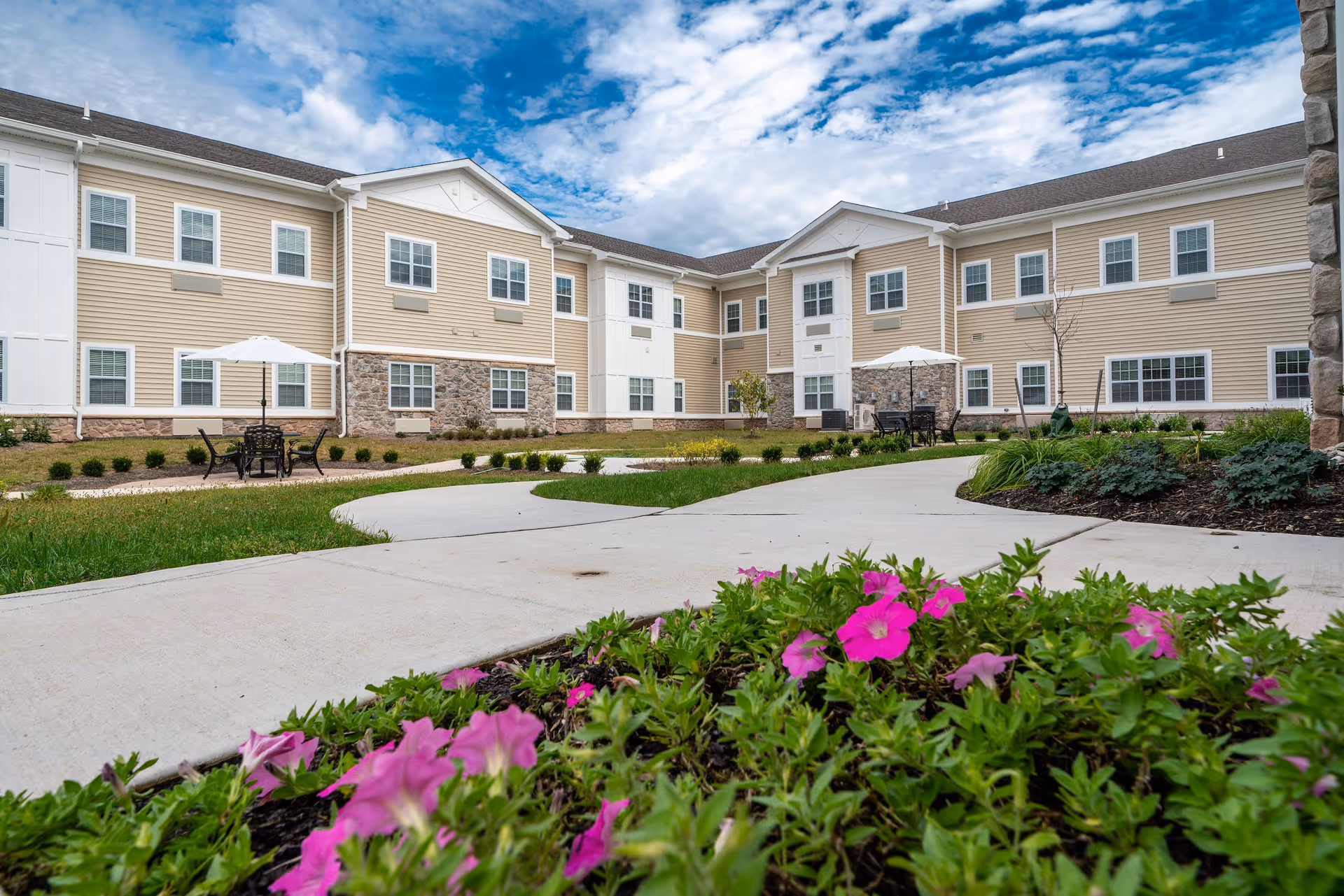 Outdoor courtyard area of Providence Place Senior Living at the Collegeville Inn with a paved walkway, green grass, flower beds with pink flowers, patio tables with umbrellas, and a beige two-story building with white trim and multiple windows under a partly cloudy sky.