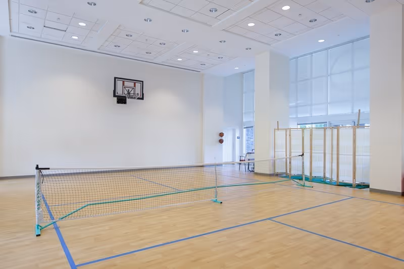 Indoor gymnasium with a wooden floor, a basketball hoop mounted on the wall, a portable net set up for a game, and large windows with blinds letting in natural light.