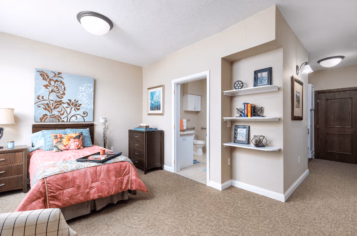 A cozy bedroom with a bed covered in a coral pink quilt and decorative pillows. There is a nightstand with a lamp and a candle, a chest of drawers, and a wall-mounted artwork above the bed. An open doorway reveals a bathroom with a toilet and white cabinets. To the right, there are three white floating shelves with decorative items and books. The room has beige walls and carpeted flooring.