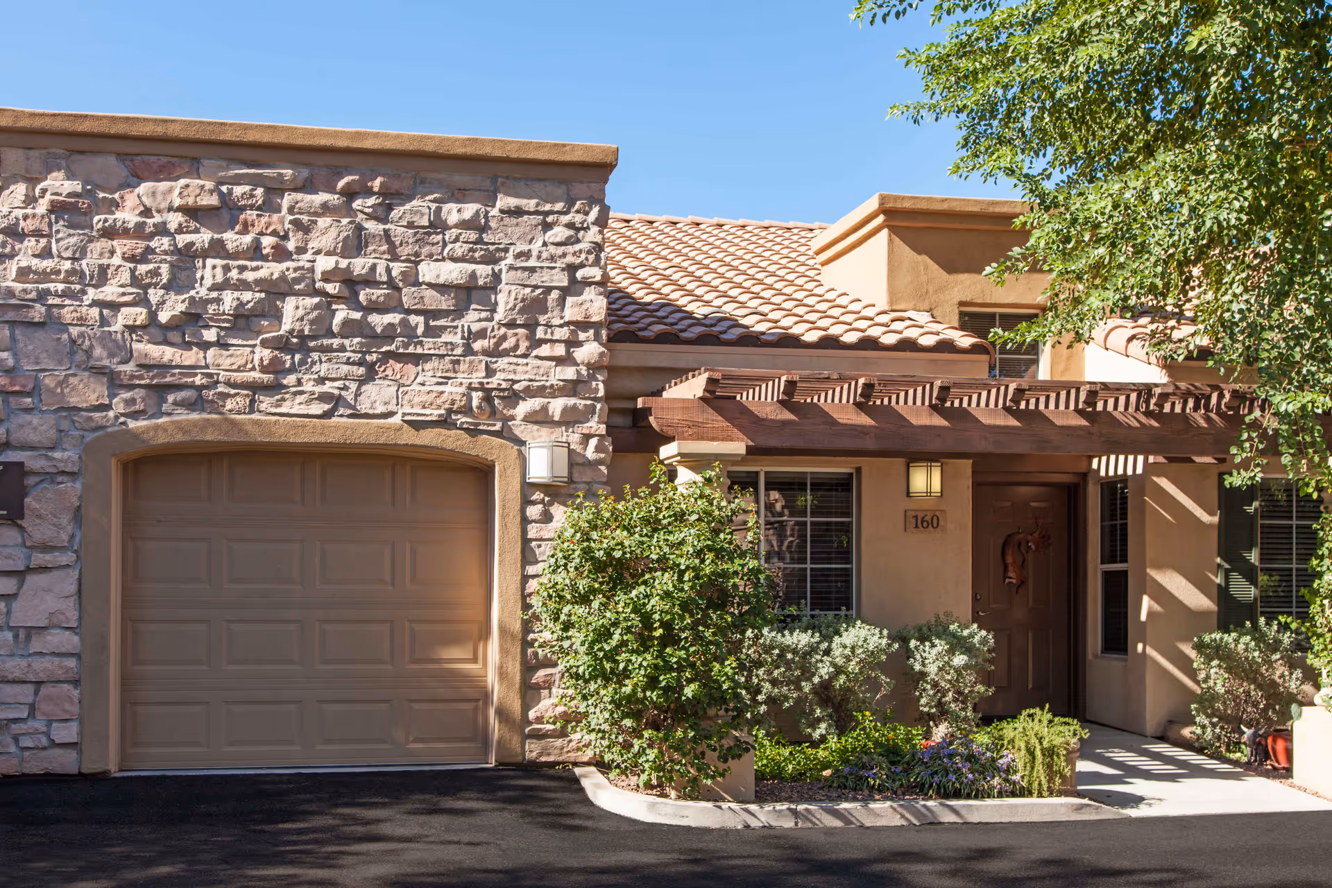 Exterior view of a residential unit at Tuscany at McCormick Ranch featuring a stone and stucco facade, a closed garage door, a wooden pergola over the entrance, and surrounding greenery including bushes and a tree.