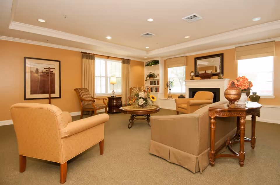 A cozy living room with beige walls and carpet, featuring several upholstered armchairs and a sofa arranged around a wooden coffee table with a floral centerpiece. There is a fireplace with a mirror above it, two windows with beige curtains, a wooden side table with decorative items, and a framed picture on the wall.