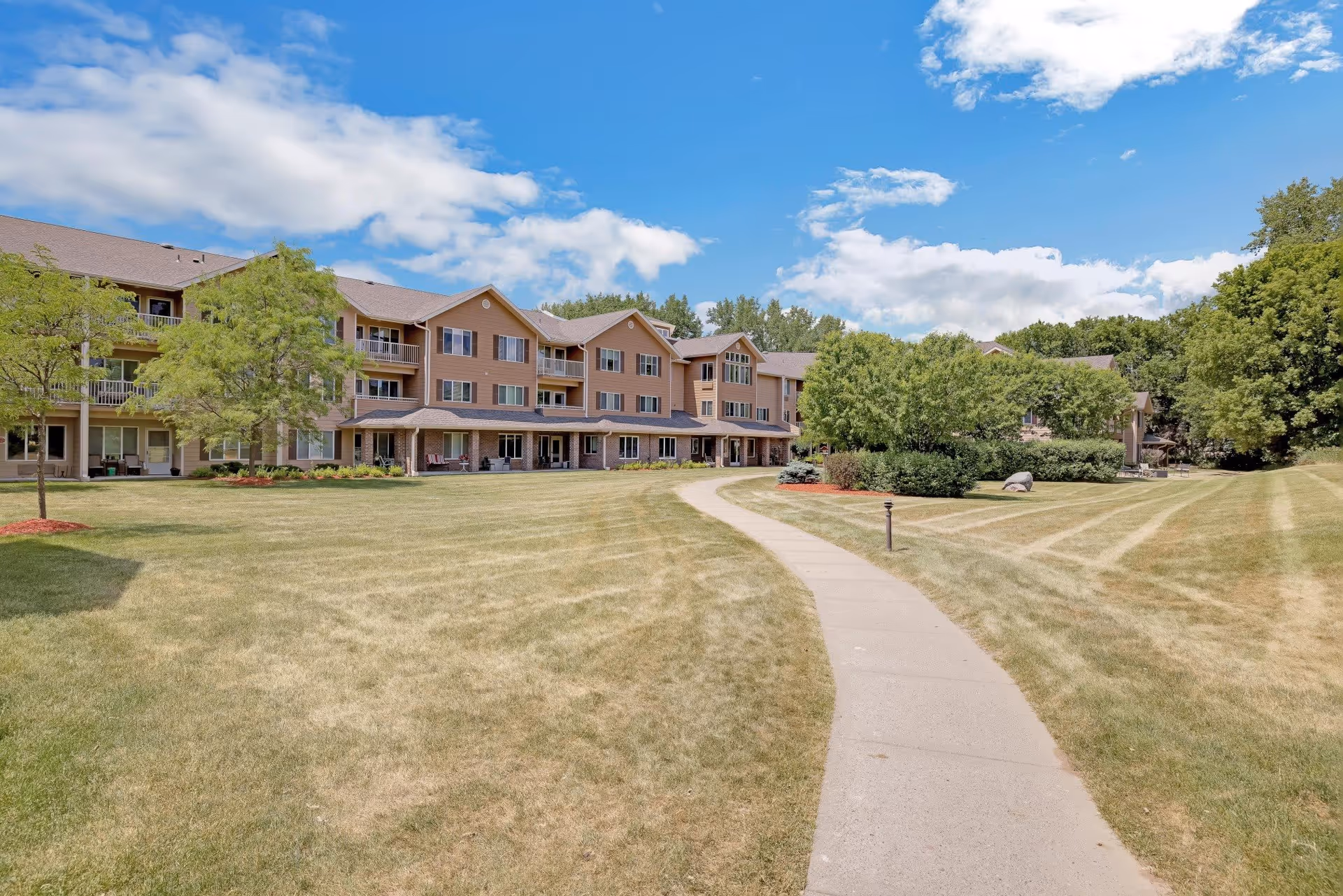 A wide view of a senior living facility building with three stories, beige siding, and multiple balconies. The building is surrounded by a large, well-maintained grassy lawn with a concrete walkway leading towards the entrance. Trees and shrubs are scattered around the lawn under a partly cloudy blue sky.