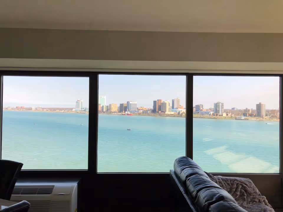 View through large windows showing a waterfront city skyline across a body of water, with part of a black leather couch and an air conditioning unit visible inside the room.