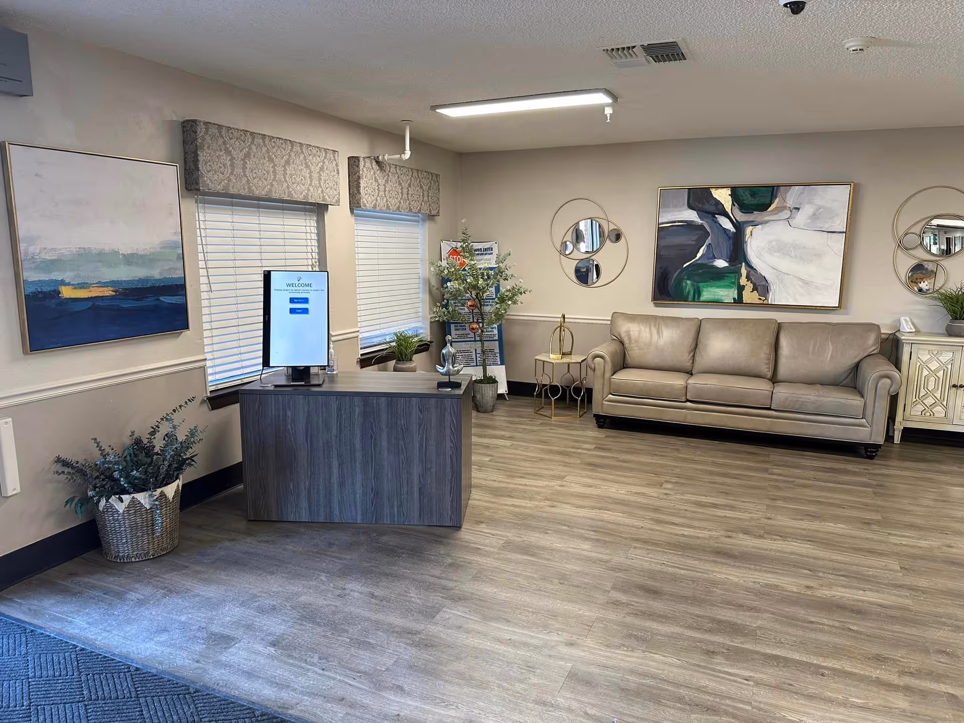 Reception area of Concho Health and Rehabilitation Center with a wooden desk featuring a computer monitor displaying a welcome screen, a beige leather sofa against the wall, decorative round mirrors, abstract paintings, potted plants, and large windows with patterned valances.