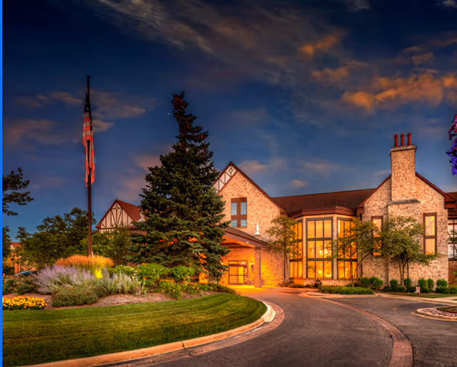 Exterior view of a large, elegant brick building at dusk with warm lights glowing from the windows. The building is surrounded by well-maintained landscaping including trees, shrubs, and flowers. An American flag is visible on a flagpole near the entrance, and a curved driveway leads up to the front door.