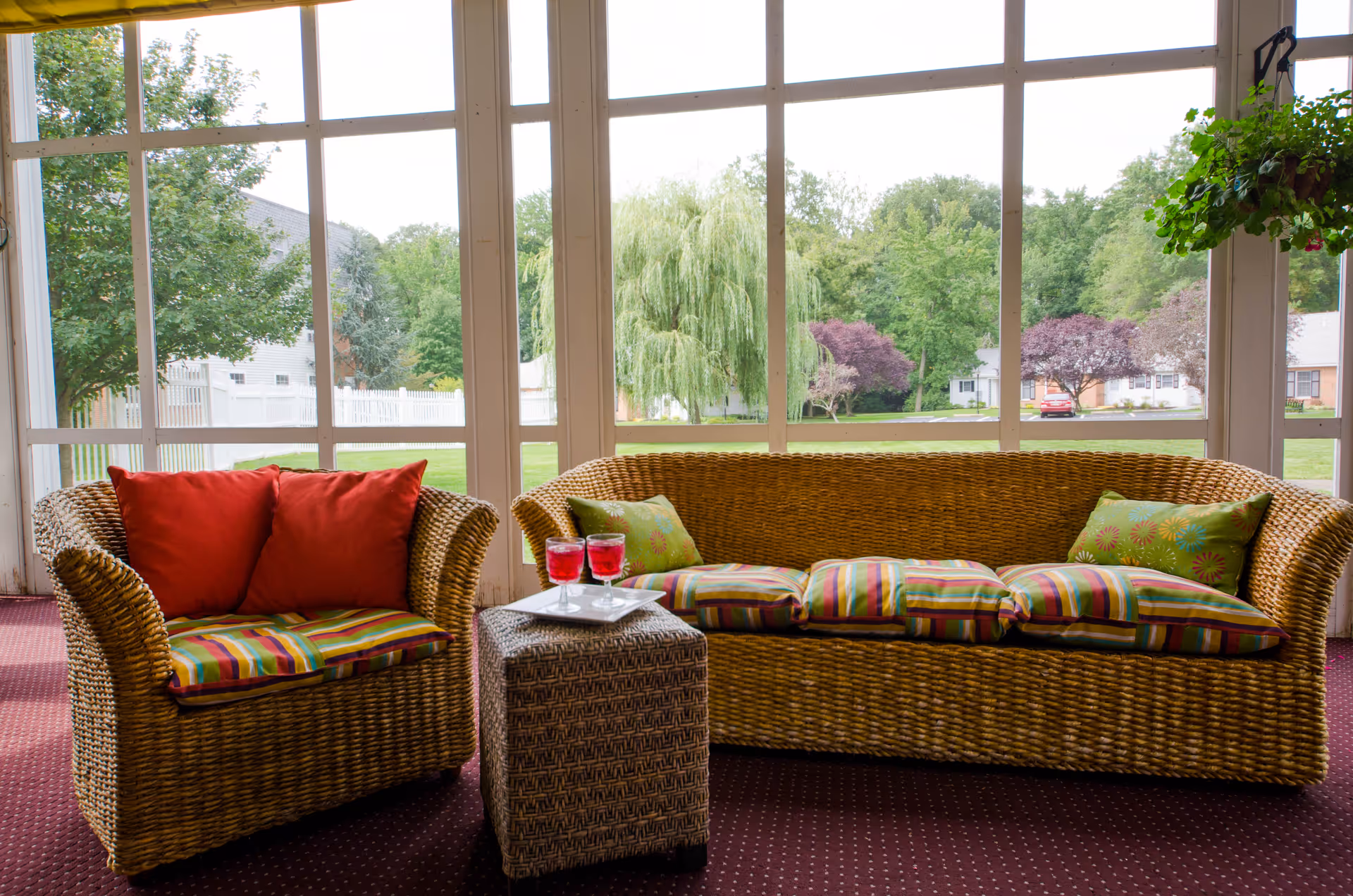 A cozy indoor seating area with wicker furniture including a loveseat and a sofa, both with colorful striped cushions and decorative pillows. A small wicker table between them holds two glasses of red beverage on a white tray. Large windows behind the furniture reveal a green outdoor landscape with trees and houses.