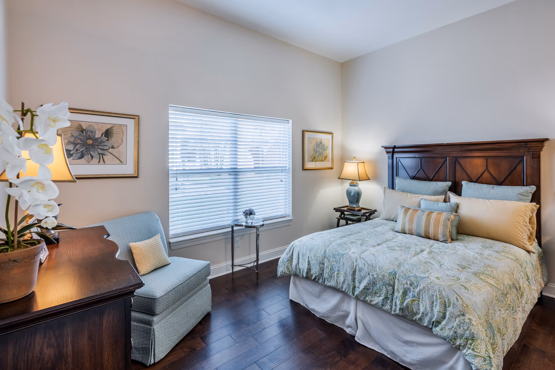 A cozy bedroom with a large wooden headboard bed adorned with multiple pillows and a patterned bedspread. There is a light blue armchair with a small pillow next to a wooden dresser with a potted orchid and a lamp. The room has hardwood floors, two framed floral artworks on the walls, and a window with white blinds letting in natural light.