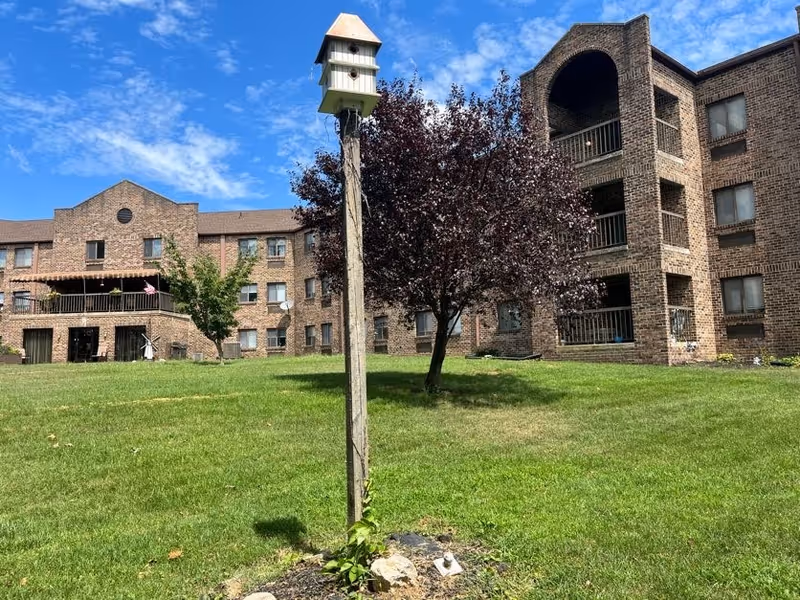 A grassy outdoor area in front of a multi-story brick building with balconies and windows. In the foreground, there is a tall wooden post with a multi-compartment birdhouse mounted on top. A tree with dark red leaves is situated near the building under a partly cloudy blue sky.