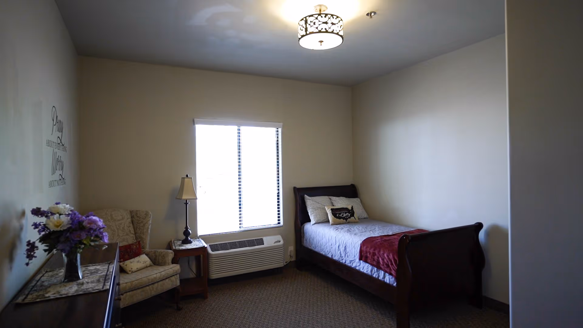 A simple bedroom with a single bed covered with a light gray quilt and a red throw blanket. There are two pillows on the bed, one with a decorative design. Next to the bed is a window with blinds and an air conditioning unit below it. To the left of the window is a wooden side table with a lamp and an upholstered armchair with a patterned fabric. A dresser with a vase of purple and white flowers is partially visible on the left side. The walls are painted light beige, and there is a ceiling light fixture with a decorative shade.