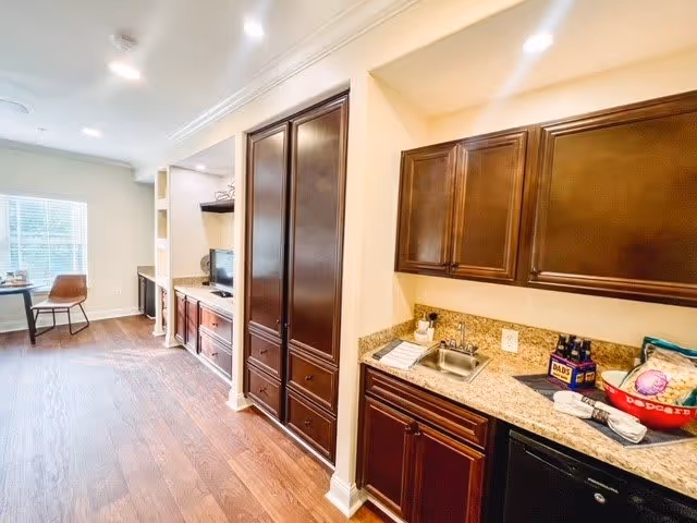 Interior view of a room with wooden flooring and dark wood cabinetry. On the right side, there is a kitchenette area with a small sink, countertop, upper and lower cabinets, a mini fridge, and various items including a bowl and bottles. On the left side, there is a built-in entertainment center with a TV and storage cabinets. A window with blinds and a chair are visible at the far end of the room.