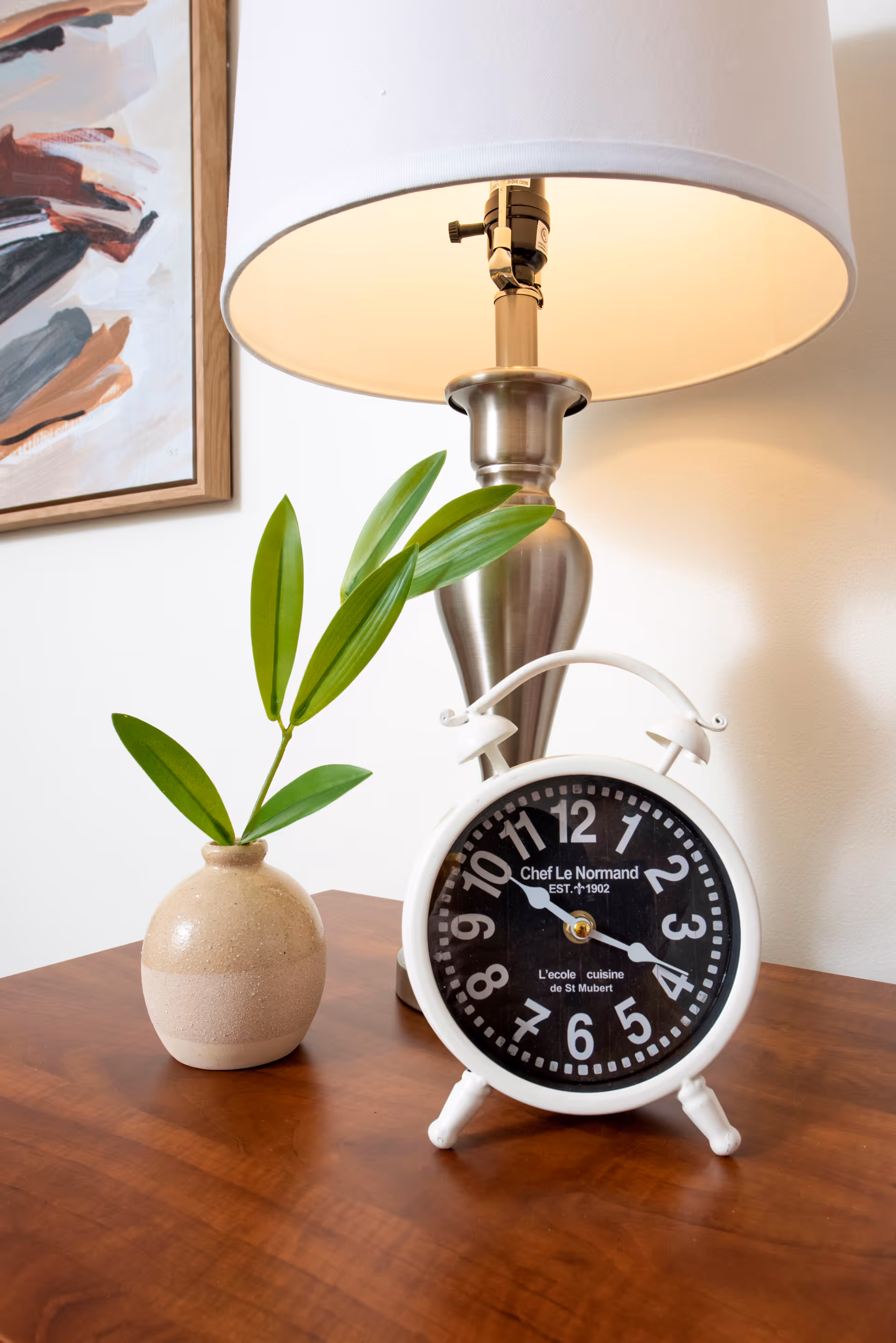 A close-up of a wooden table with a small beige vase holding green leaves, a silver table lamp with a white lampshade, and a white vintage-style alarm clock with a black face displaying the time 4:20. A framed abstract painting is partially visible on the wall behind.
