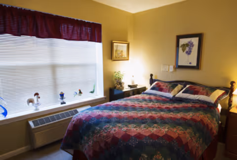 A cozy bedroom with a double bed covered in a colorful quilt. The room has beige walls with two framed pictures hanging above the bed and a window with closed blinds and a red valance. There is a small black nightstand with a lamp and decorative items next to the bed, and a wooden dresser on the right side.