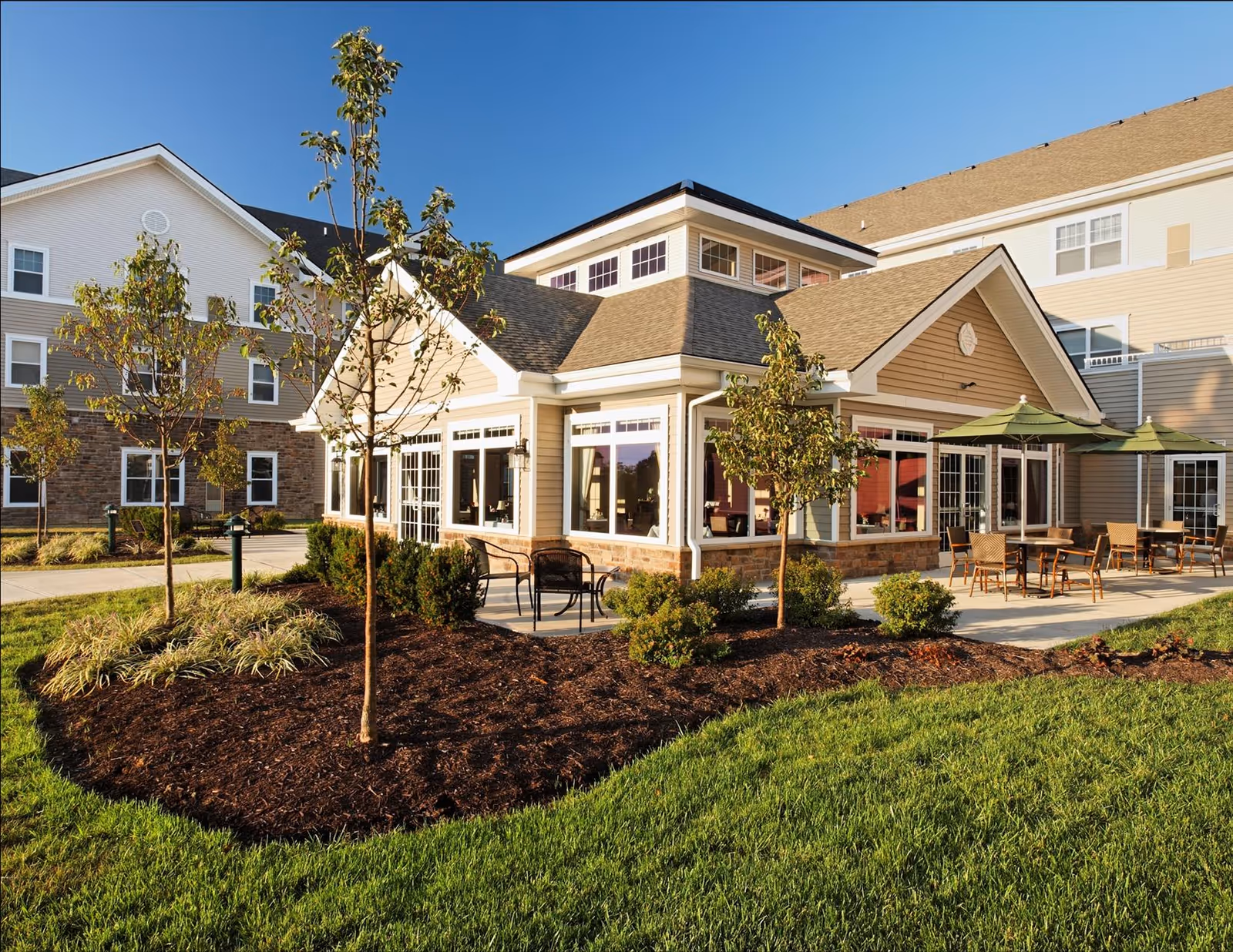 Exterior view of a senior living facility building with beige siding and stone accents. The building has large windows and a patio area with tables, chairs, and green umbrellas. There are small trees and landscaped bushes in the foreground with a well-maintained lawn under a clear blue sky.