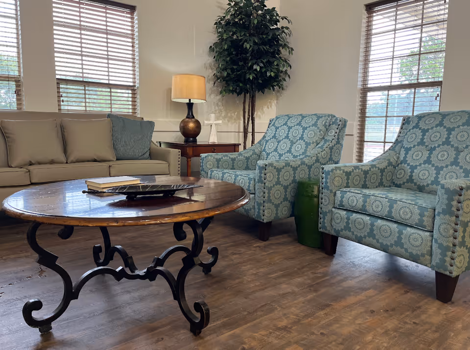 A seating area with a sofa, two patterned armchairs, a wooden coffee table, a lamp and a potted tree in a bright living room.
