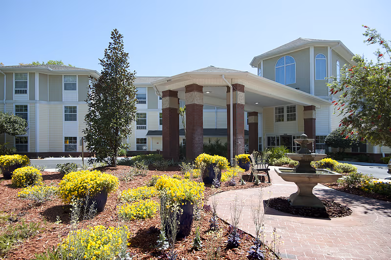 Exterior view of Allegro Senior Living in Tallahassee, FL, showing the main entrance with a covered drop-off area supported by brick columns. The foreground features a landscaped garden with yellow flowers, a stone fountain, and a curved brick walkway leading to the entrance. The building is multi-story with light-colored siding and large windows.