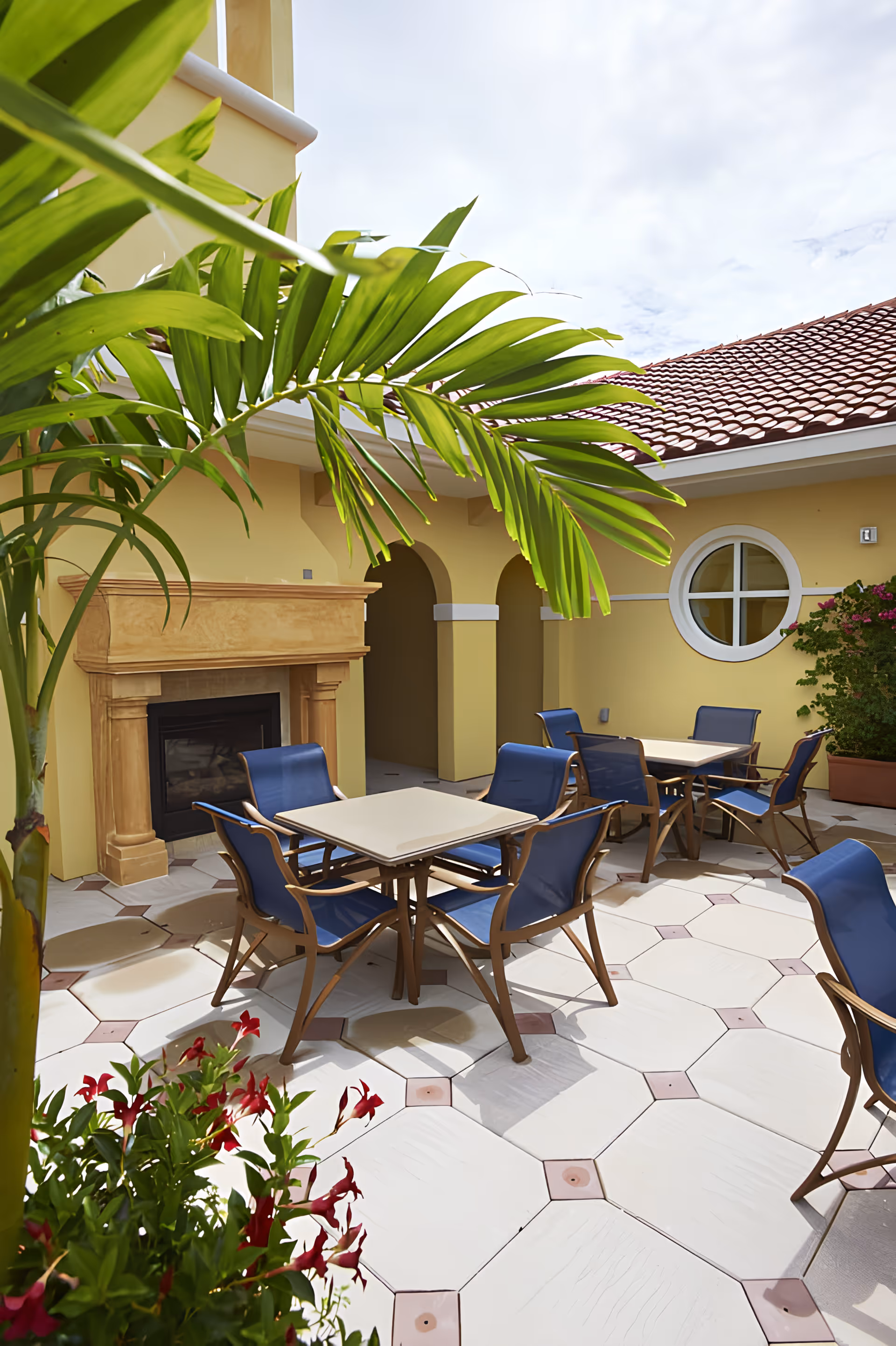 Sunlit courtyard with patio tables and blue chairs, a fireplace, and tropical plants against yellow walls and a tiled roof.