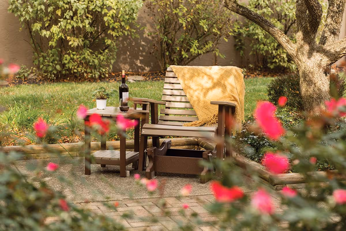 A wooden outdoor chair with a yellow blanket draped over the backrest sits next to a small wooden table holding a bottle of wine, a wine glass, and a small potted plant. The setting is a garden with green grass, bushes, a tree, and pink flowers in the foreground.
