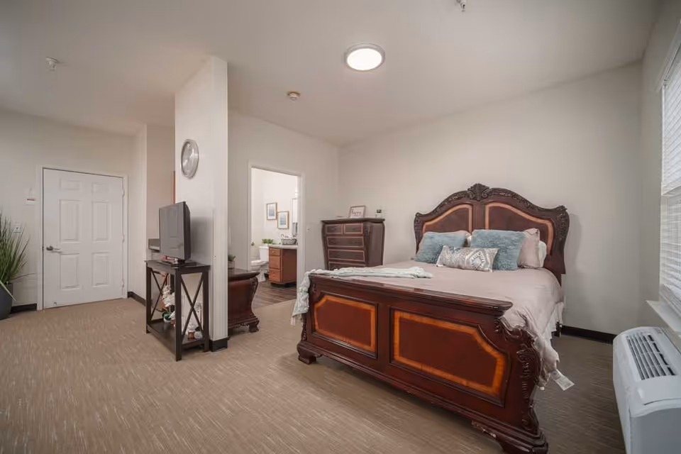 A bedroom in a senior living facility featuring a large wooden bed with an ornate headboard and footboard, decorated with pillows and a blanket. There is a wooden dresser against the wall and a TV on a stand near the entrance. A doorway leads to a bathroom with a visible toilet and sink. The room has beige walls, carpeted floor, and a window with blinds on the right side.