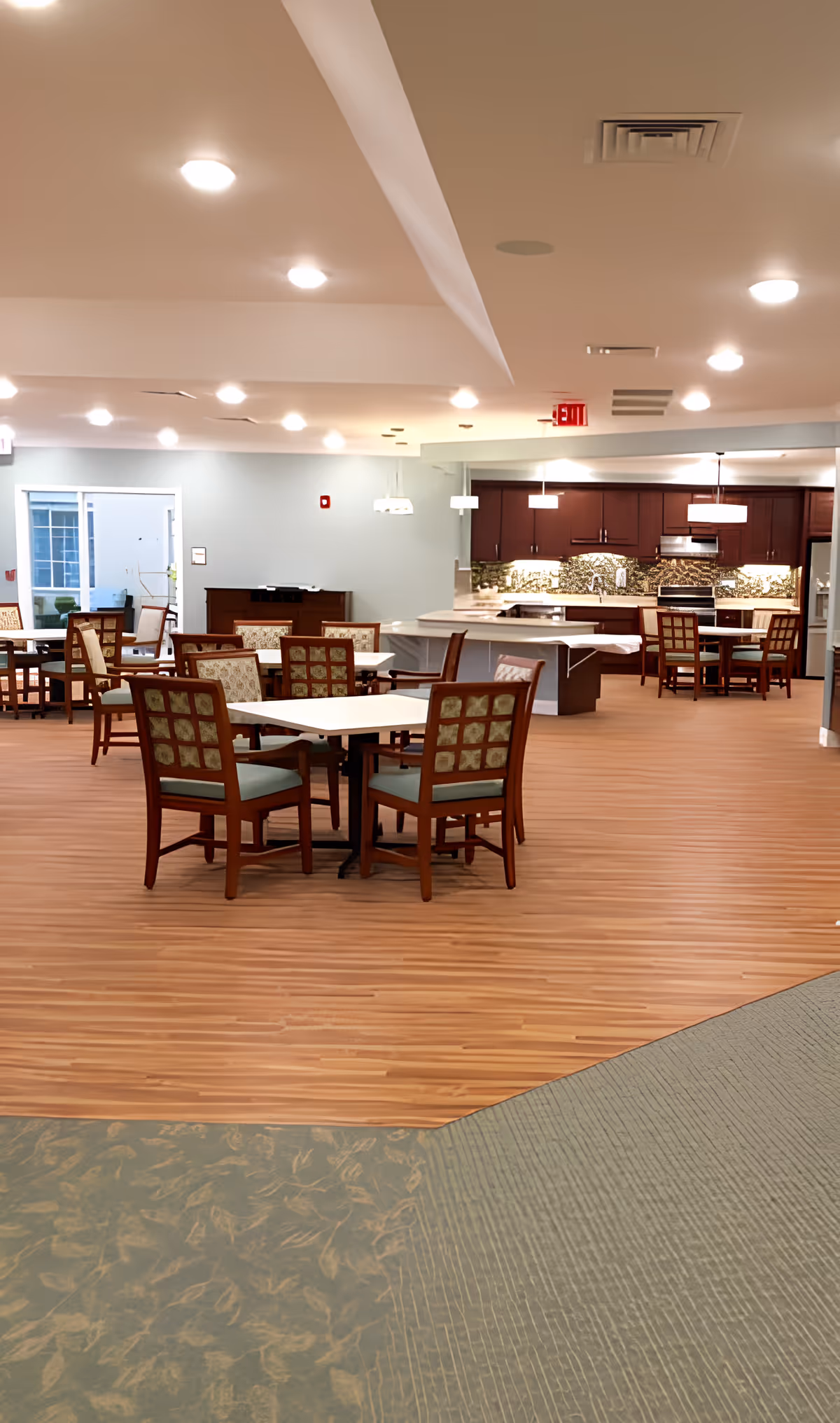 A spacious dining area in The Ganzhorn Suites featuring multiple tables with wooden chairs upholstered in patterned fabric. The room has wood flooring and a section of carpet with a leaf pattern. In the background, there is a kitchen area with dark wood cabinets, a stove, and pendant lighting. The ceiling is equipped with recessed lights and an exit sign is visible.