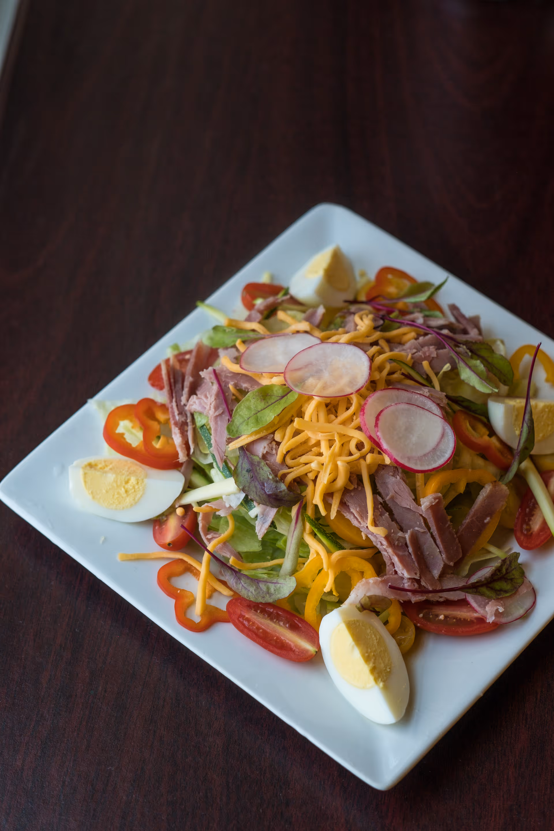 A square white plate with a fresh salad consisting of sliced hard-boiled eggs, shredded cheddar cheese, sliced radishes, cherry tomato halves, sliced bell peppers, leafy greens, and strips of ham on a dark wooden table.