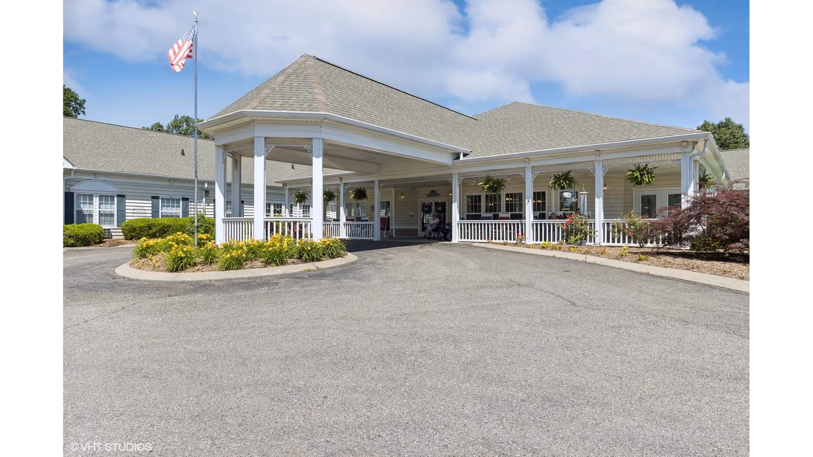 Exterior view of The Bungalows at Mayfield senior living facility showing a covered entrance with white pillars, a circular driveway, landscaped plants, an American flag on a flagpole, and a clear blue sky with some clouds.
