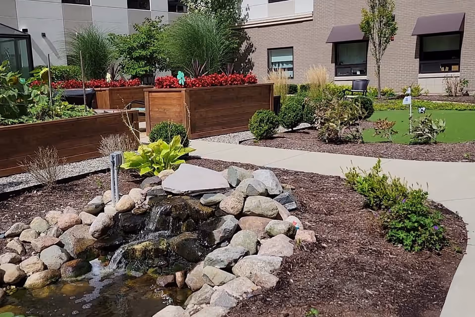 Outdoor garden area at Willowbrook Hills featuring a small rock waterfall pond, raised wooden planter boxes with red flowers, various shrubs and plants, a paved walkway, and a building with windows in the background.