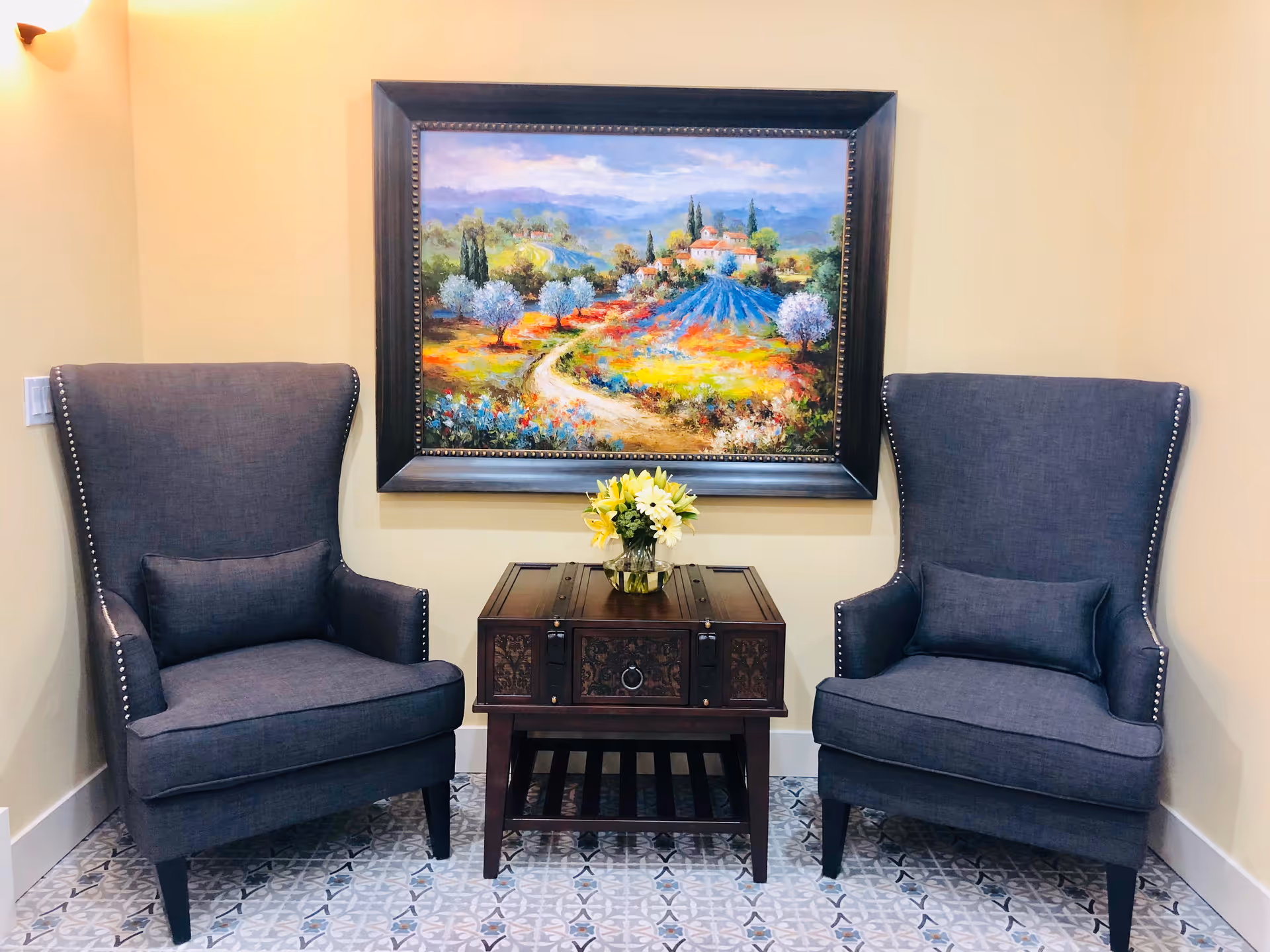 Two dark gray upholstered armchairs with nailhead trim are positioned on either side of a small wooden table with intricate carvings. A vase with yellow and white flowers sits on the table. Above the table hangs a large framed painting depicting a colorful countryside landscape with trees, flowers, and a winding path leading to a house.