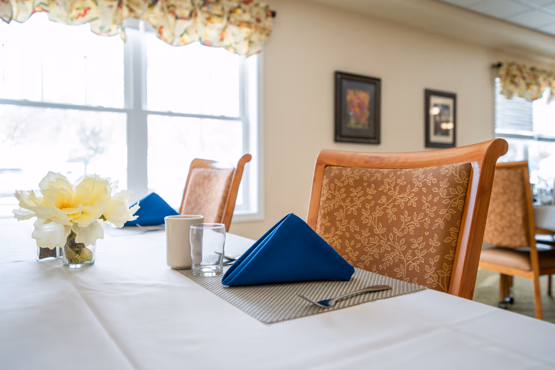 A bright dining room table set with blue folded napkins, a glass and cup, a small floral centerpiece, and patterned wooden chairs by large windows.