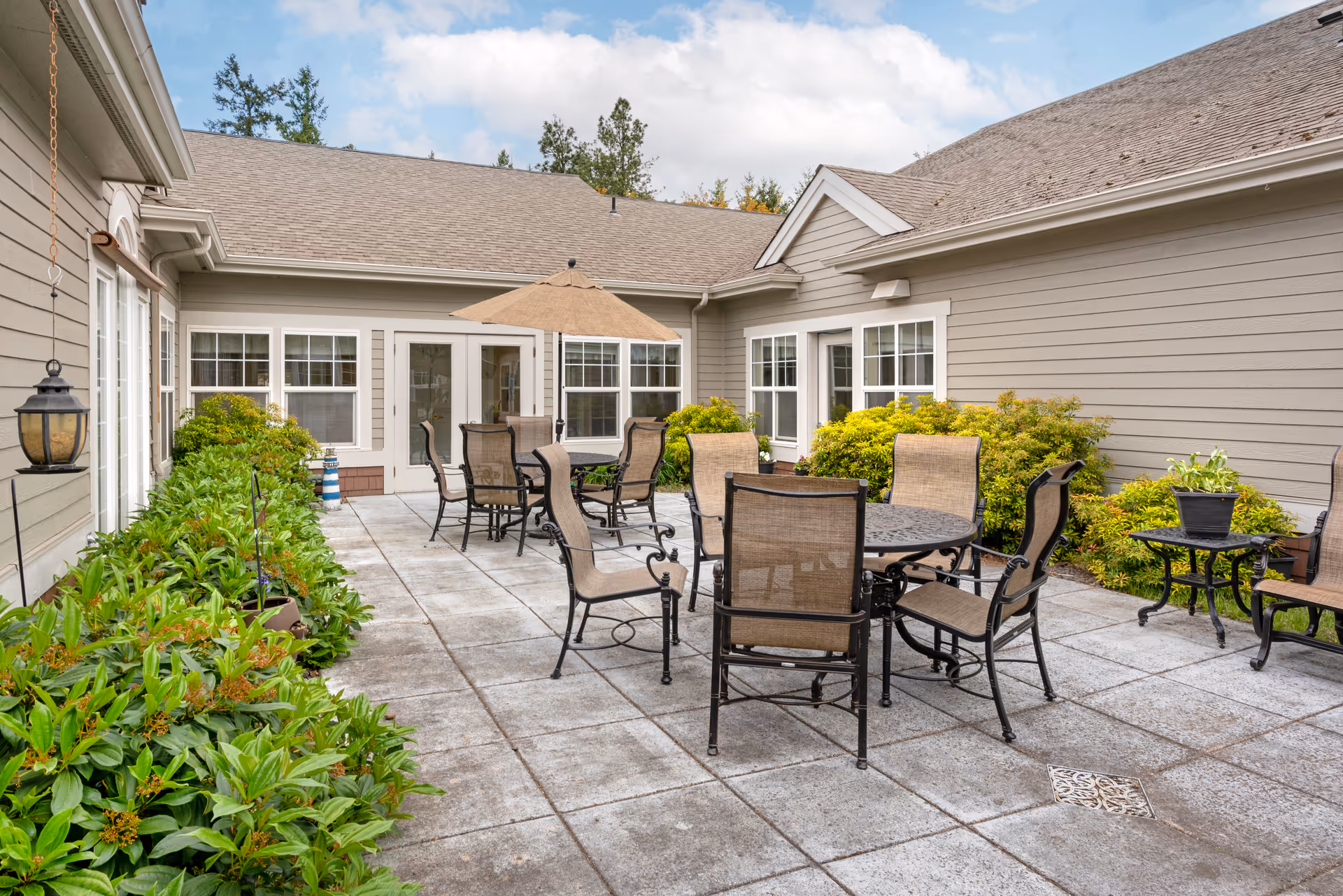 Outdoor patio area at Brookdale Salem with multiple round metal tables and chairs, a large umbrella, surrounded by green shrubs and beige siding buildings under a partly cloudy sky.