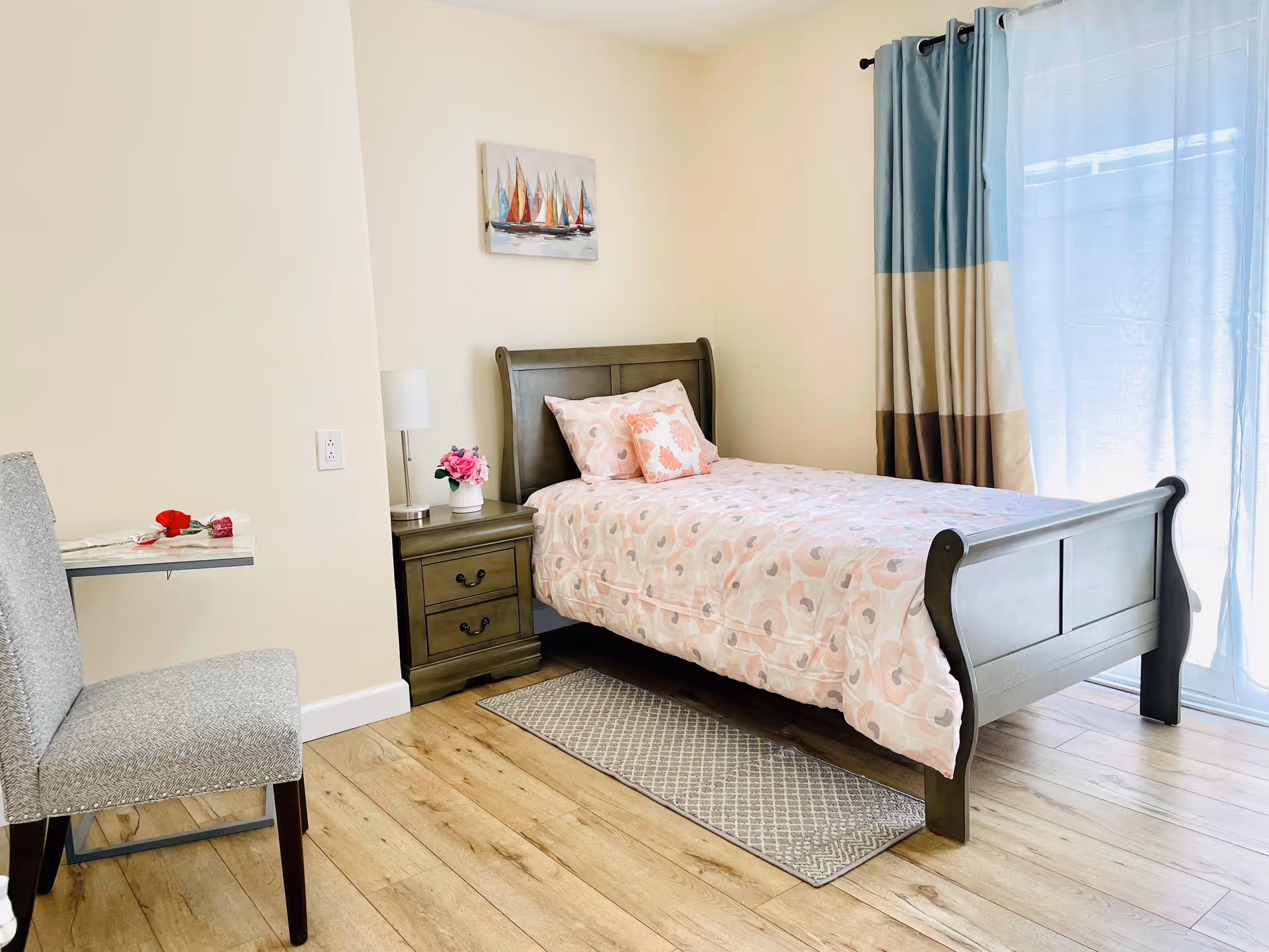 A bright and cozy bedroom with a single wooden bed covered in a pink floral bedspread and matching pillows. Next to the bed is a wooden nightstand with a small lamp and a vase of pink flowers. A gray upholstered chair is positioned near a small wall-mounted table with a red rose on it. The room has light-colored walls, wood flooring, a small patterned rug, and a window with blue, beige, and brown curtains letting in natural light. A painting of colorful sailboats hangs on the wall above the bed.