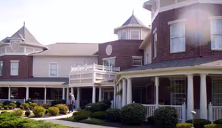Exterior view of a large brick and white senior living facility building with multiple windows, a porch with white columns, and well-maintained bushes and lawn in front.