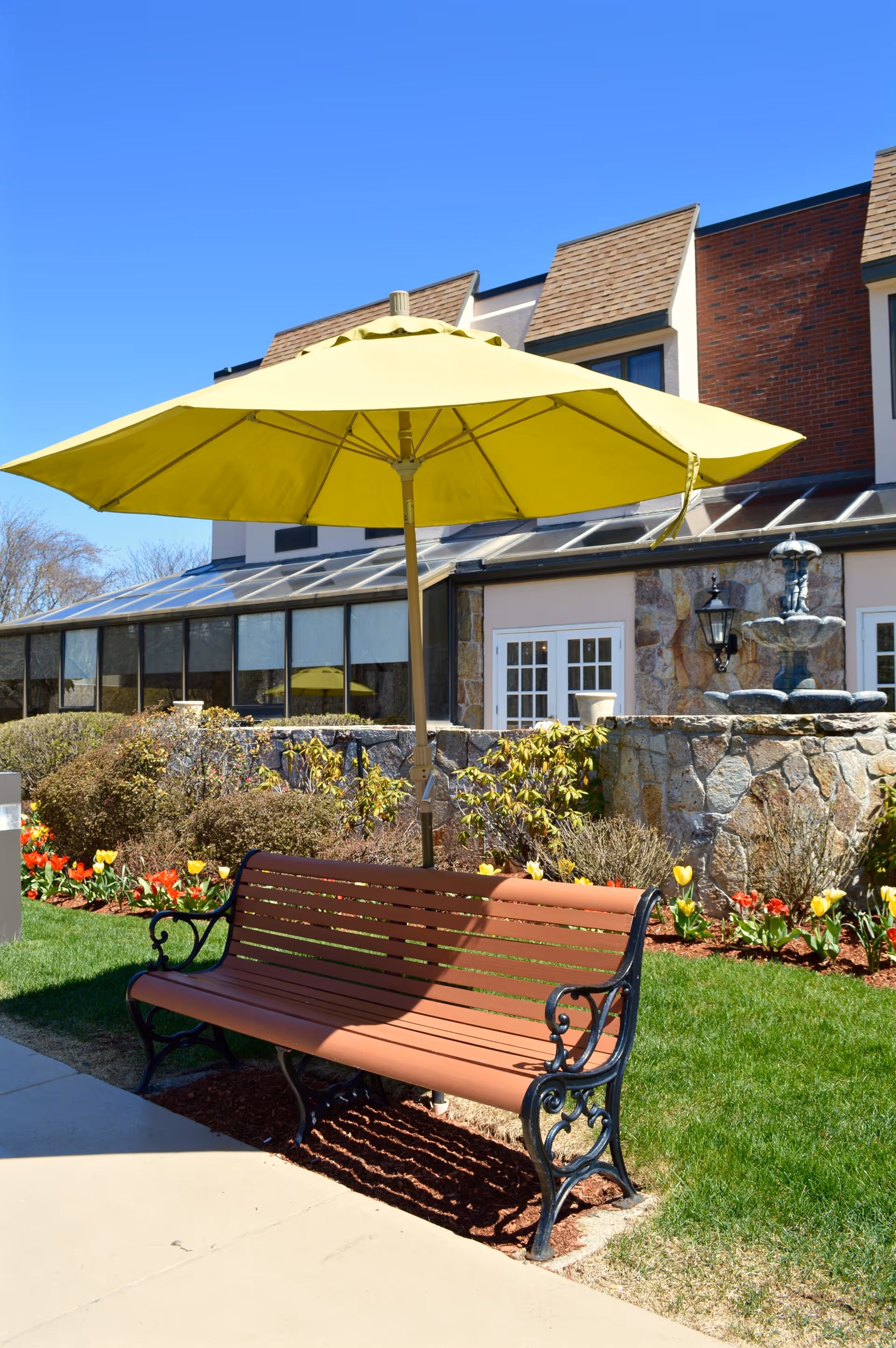 A wooden bench with black metal armrests and legs sits on a sidewalk next to a garden with colorful tulips and bushes. A large yellow umbrella is positioned above the bench providing shade. Behind the garden is a stone wall with a water fountain and a building with windows and a sloped roof under a clear blue sky.