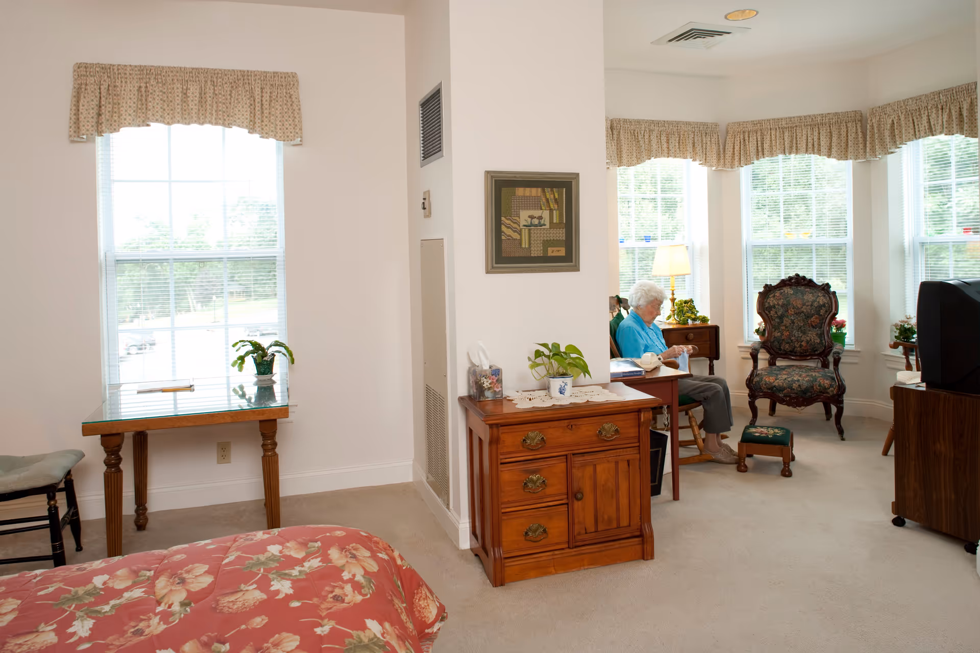 A cozy room in a senior living facility with a floral bedspread in the foreground, a wooden table with a plant near a window, and an elderly woman sitting on a chair near a corner with large windows and floral curtains. The room has beige carpet and wooden furniture including a small dresser and a TV stand.