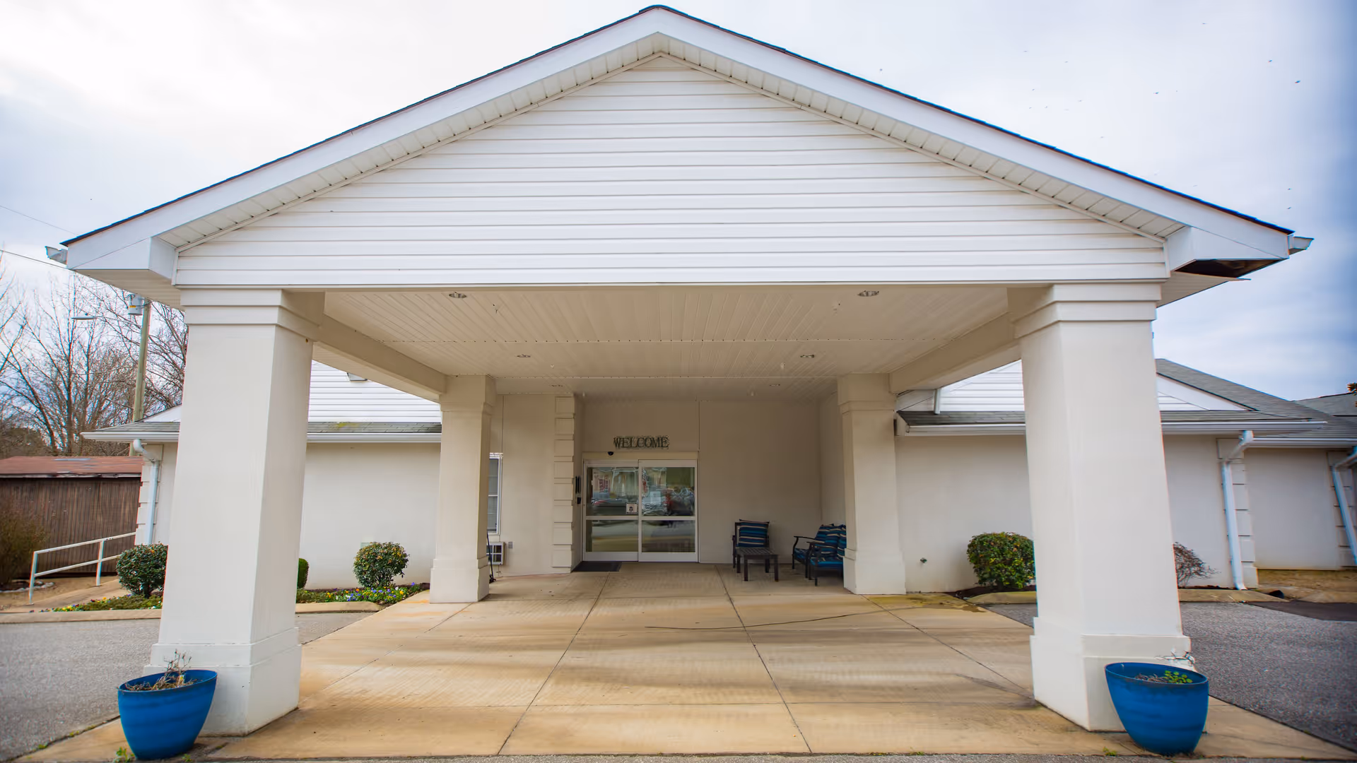Entrance of a single-story building with a covered driveway supported by four large white columns. There are two blue planters with small plants on either side of the driveway. The building has a white exterior with a peaked roof, and a glass door entrance with a 'WELCOME' sign above it. Some chairs are placed near the entrance.