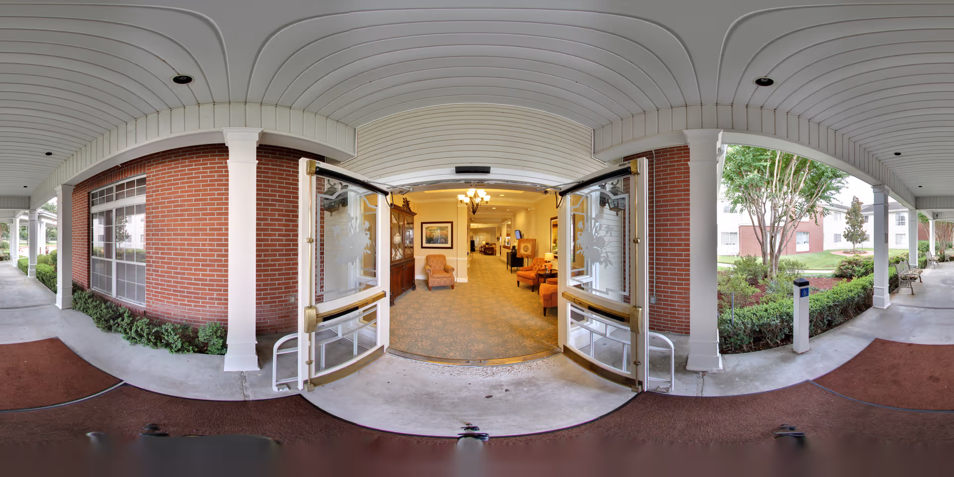 Wide-angle view of the entrance to a senior living facility showing double glass doors open to a carpeted interior lounge area with chairs, a chandelier, and framed artwork. The exterior features a covered walkway with white columns, red brick walls, and landscaped greenery including bushes and trees.