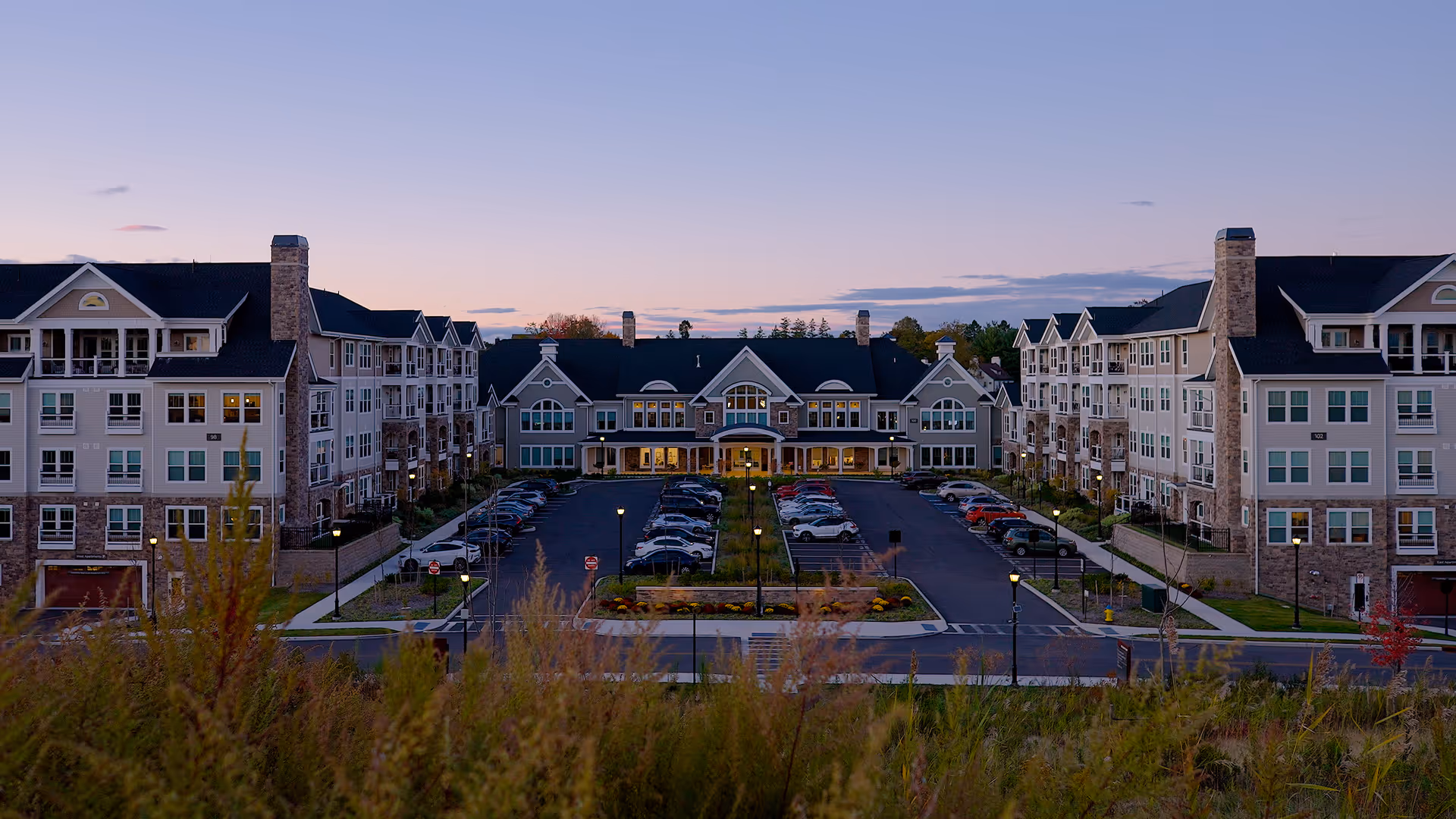 Exterior view of Broadview Senior Living at Purchase College during dusk, showing a large, multi-story building with stone and siding facade, multiple windows, and a central entrance with a covered drop-off area. There is a parking lot with cars in front of the building and landscaped greenery in the foreground.