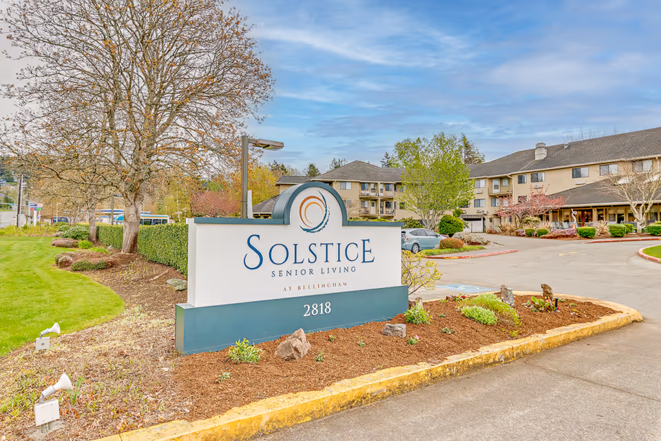 Exterior view of Solstice Senior Living at Bellingham facility with a large sign displaying the name and address 2818. The building is a multi-story structure surrounded by landscaped greenery, trees, and a driveway with parked cars under a partly cloudy sky.