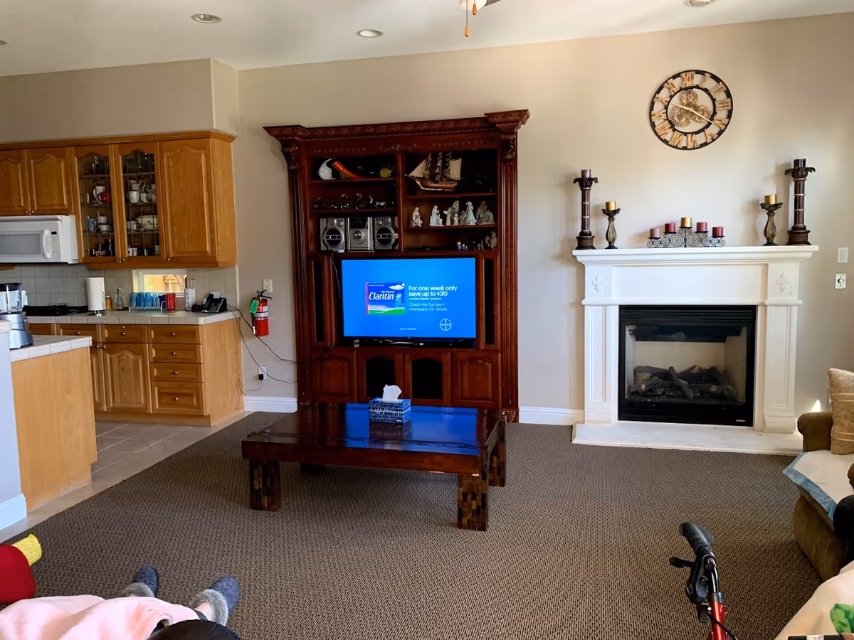 Living room with a wooden entertainment center and TV, a white fireplace, coffee table, and open view into a kitchen area.