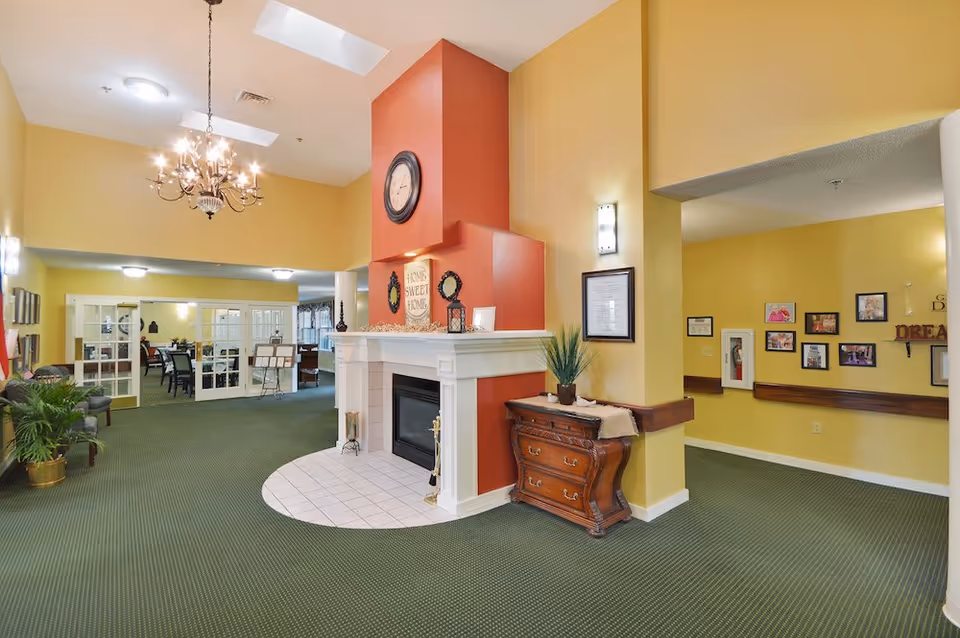 Interior view of a senior living facility common area with a green carpet, yellow walls, and a white fireplace with a red chimney. Above the fireplace is a round clock and a sign that reads 'Home Sweet Home.' There is a wooden chest of drawers next to the fireplace, a chandelier hanging from the ceiling, and framed pictures on the walls. In the background, there are glass double doors leading to a dining area with tables and chairs.