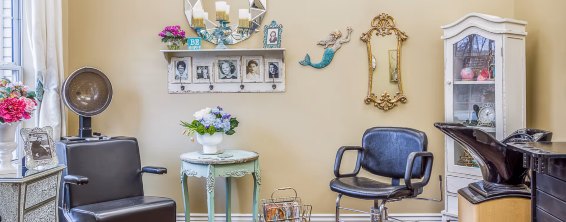 A cozy salon area with two black salon chairs, a vintage hair dryer, a small round table with a flower arrangement, decorative wall art including framed photos, a mirror, and a mermaid figure, and a white cabinet with glass doors displaying decorative items.