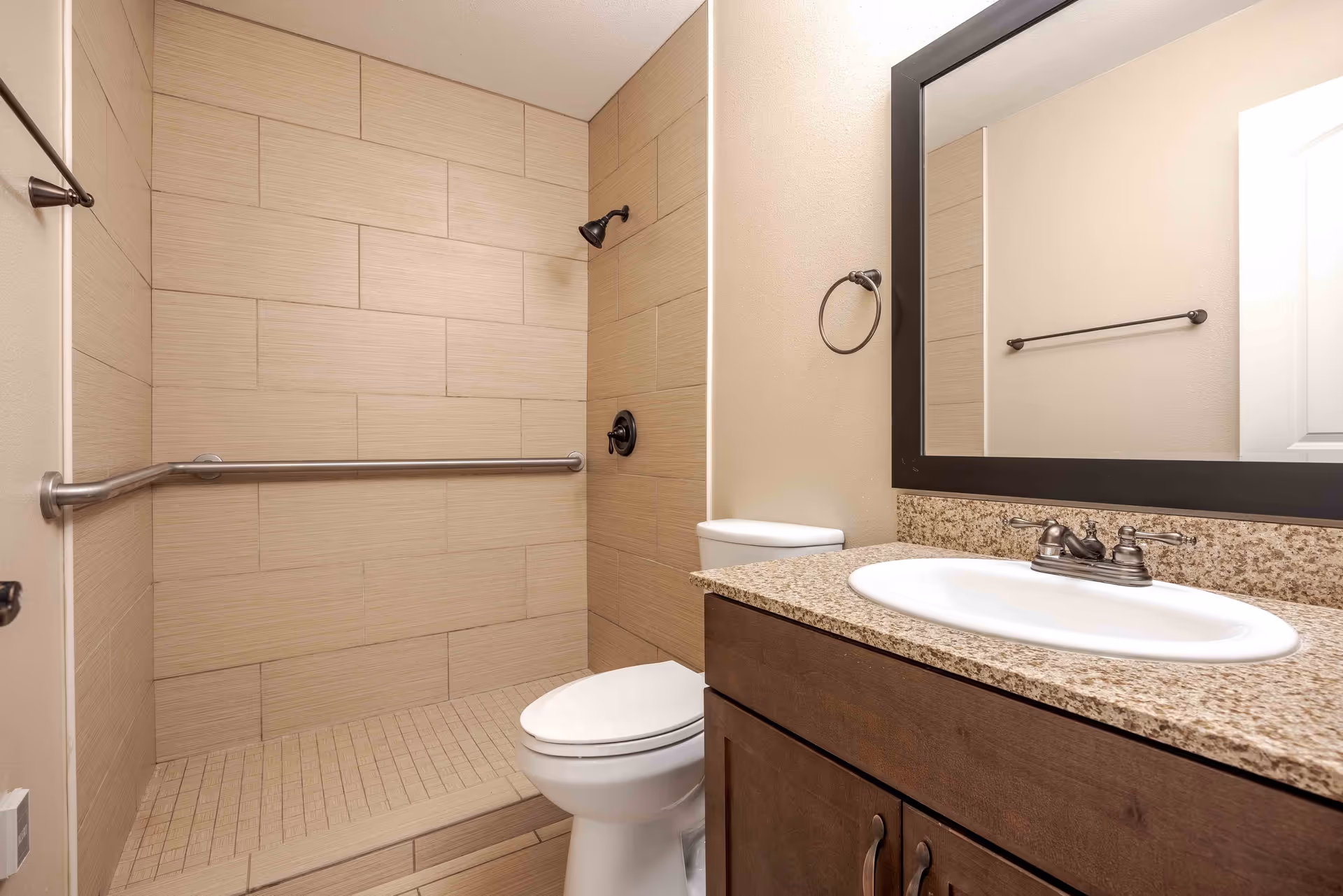 Accessible bathroom with a tiled walk-in shower and grab bar, toilet, sink with granite countertop, and wall mirror.